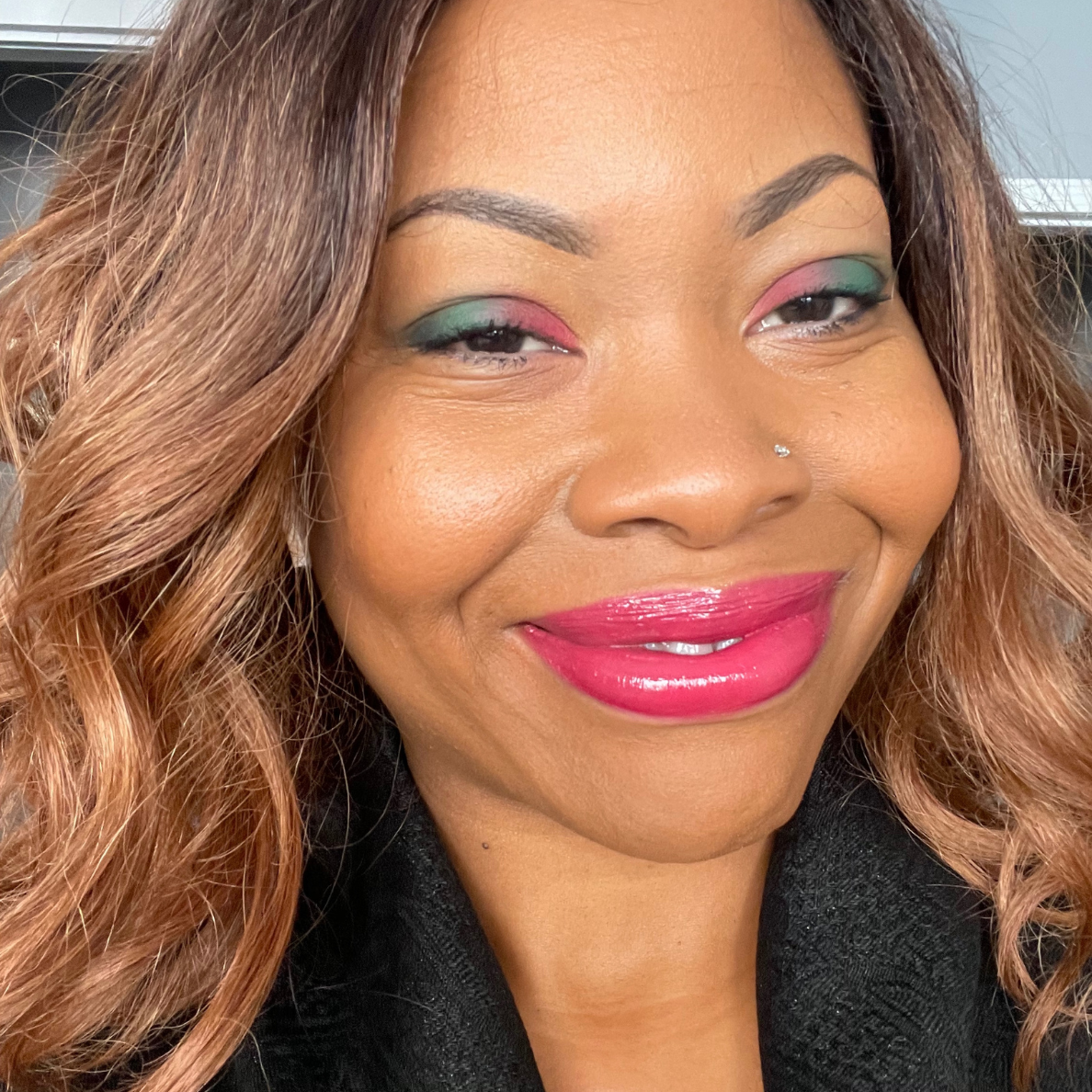 Close-up portrait of a woman with wavy brown hair, wearing colorful eye makeup featuring green and pink shades, and glossy pink lips. She is smiling and has a small nose piercing.